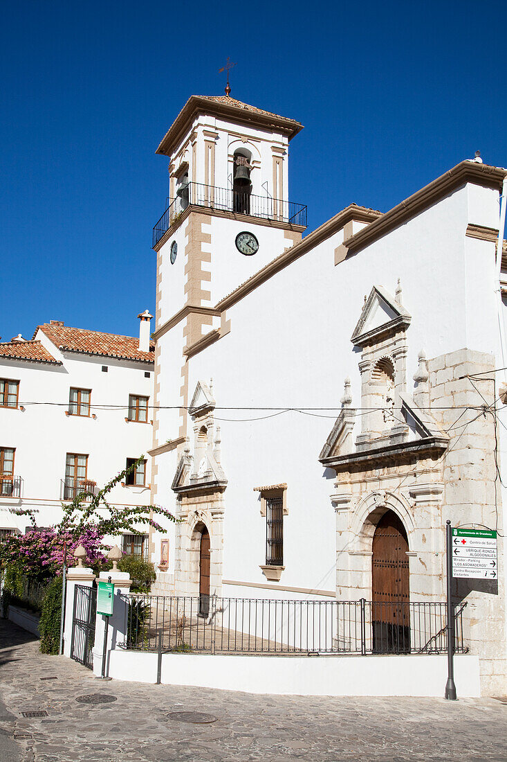 Weißes Kirchengebäude mit einer Uhr und einem Kreuz auf einem Turm vor blauem Himmel; Grazalema Andalusien Spanien