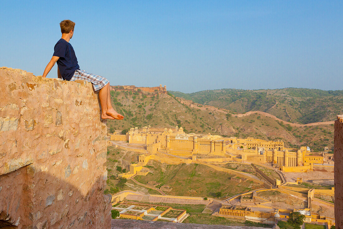 Ein junger Mann sitzt barfuß auf der Spitze einer Mauer und blickt auf das Amer Fort; Jaipur Rajasthan Indien