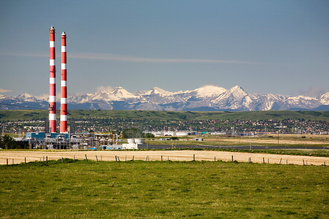 Two Red And White Stripped Smoke Stacks Of A Gas Plant With Snow Peaked Mountains In The Background And Blue Sky; Alberta Canada