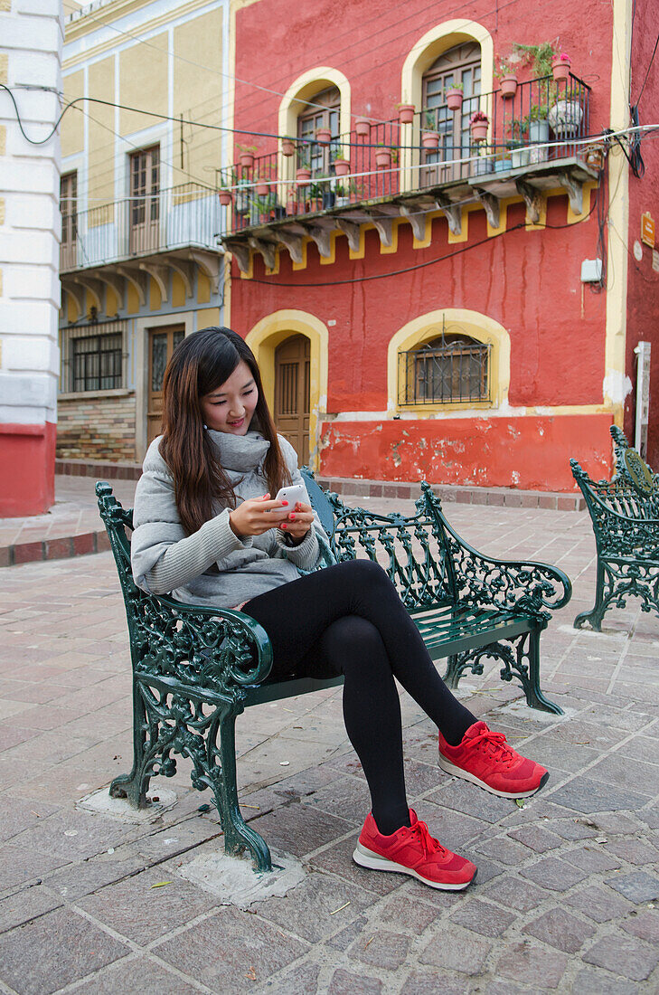 Junge Koreanerin sitzt auf einer Parkbank in einer alten spanischen Stadt und schaut auf ihr Handy; Guanajuato, Guanajuato State, Mexiko