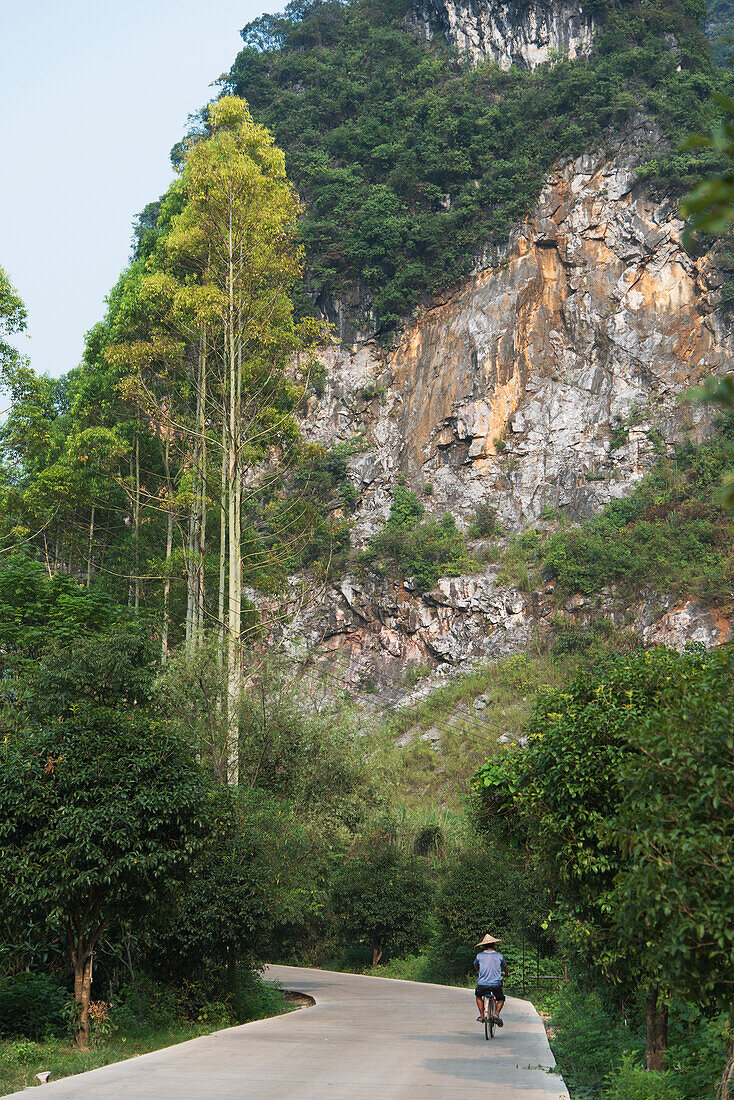 A Man Rides A Bicycle Down A Road Beside A Steep Mountainside; Guilin, Guangxi, China