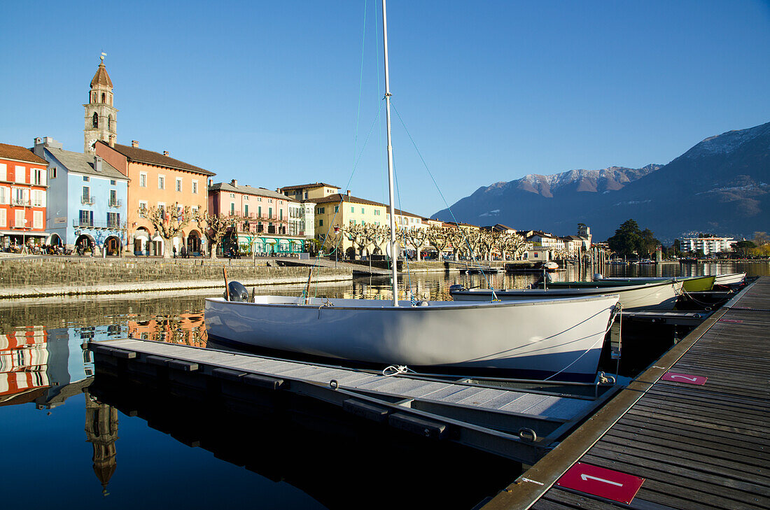 Boote ankern entlang der Docks im Lago Maggiore mit Gebäuden und den Schweizer Alpen im Hintergrund; Ascona, Tessin, Schweiz