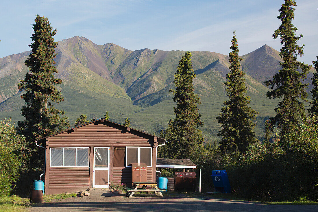Ein kleines Haus, umgeben von großen Bäumen und Bergen im Hintergrund; Yukon, Kanada