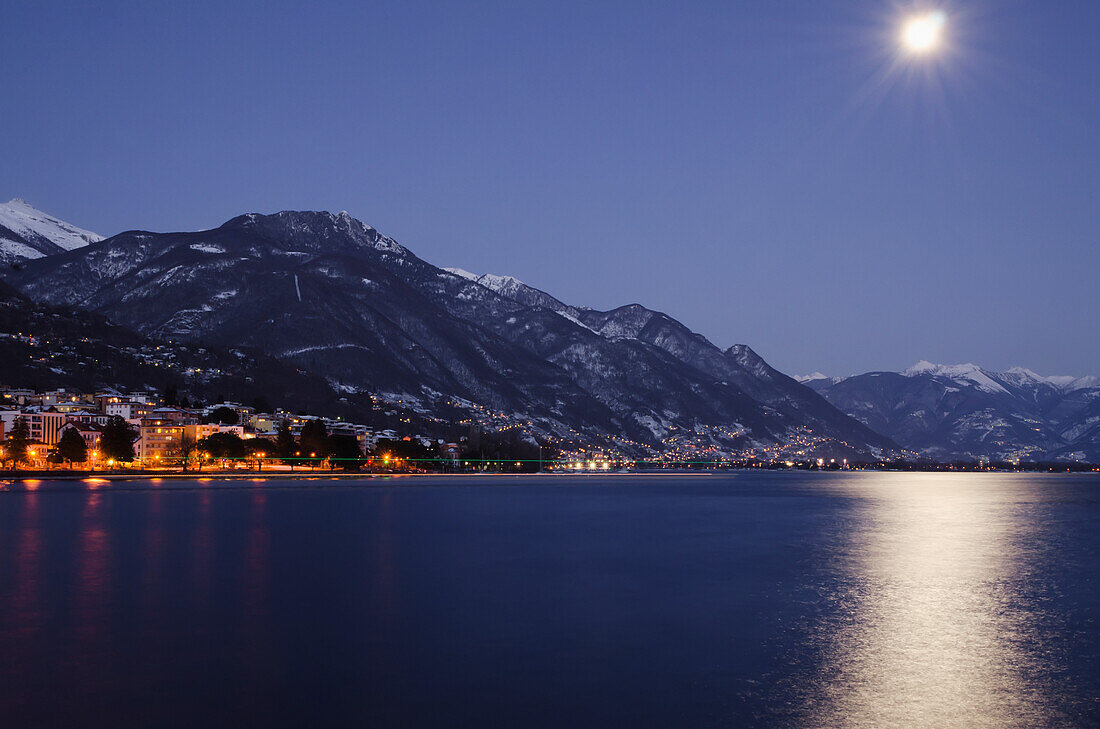 Mondlicht spiegelt sich auf dem Lago Maggiore mit beleuchteten Gebäuden entlang der Uferlinie; Locarno, Tessin, Schweiz