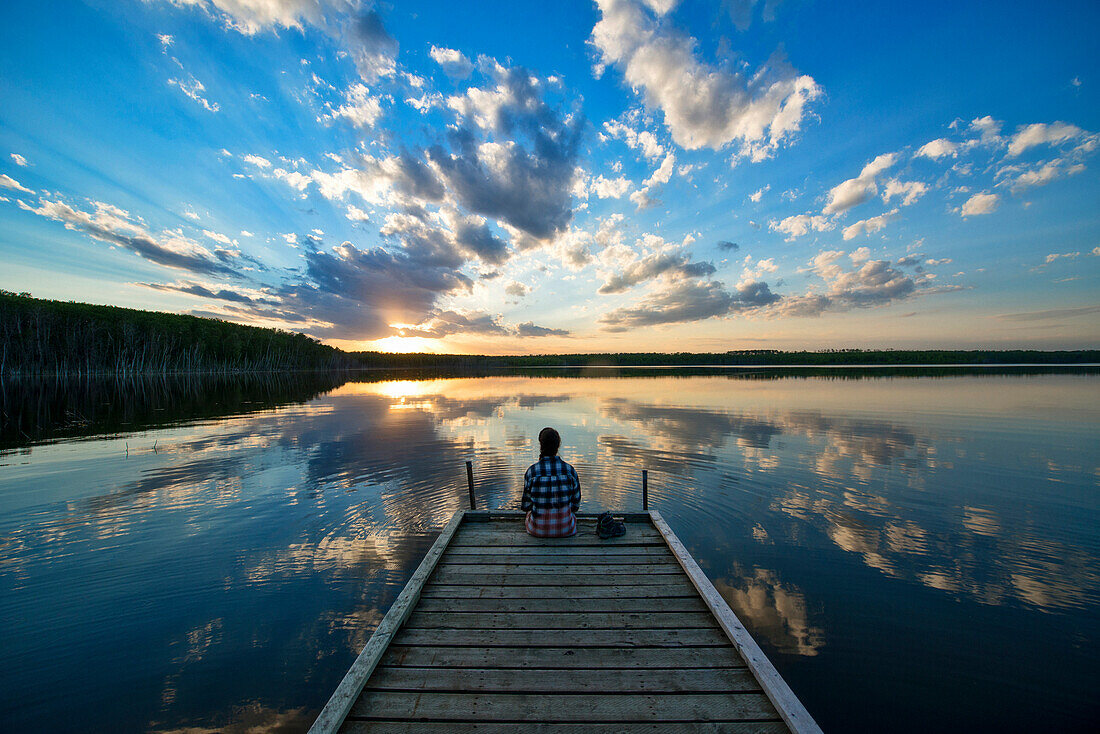 Wanderer sitzt auf Dock am ruhigen See bei Sonnenuntergang, Saskatchewan, Kanada