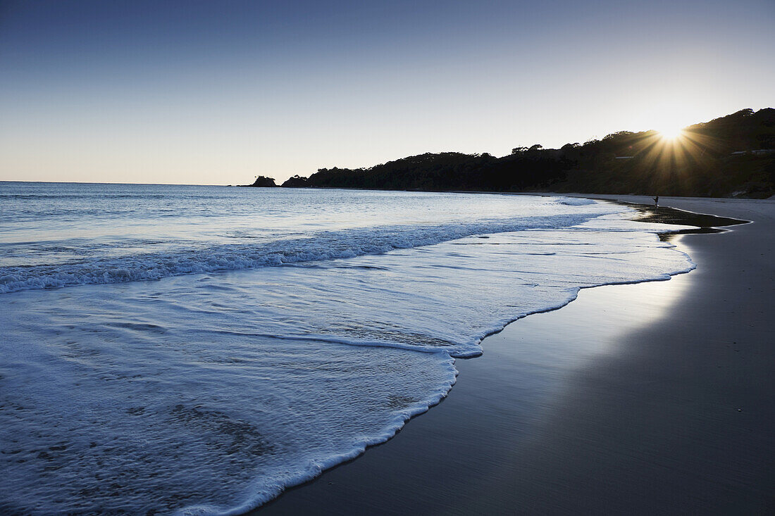 Brandung an einem Strand mit Sonnenschein über einem Hügel in Byron Bay in New South Wales, Australien