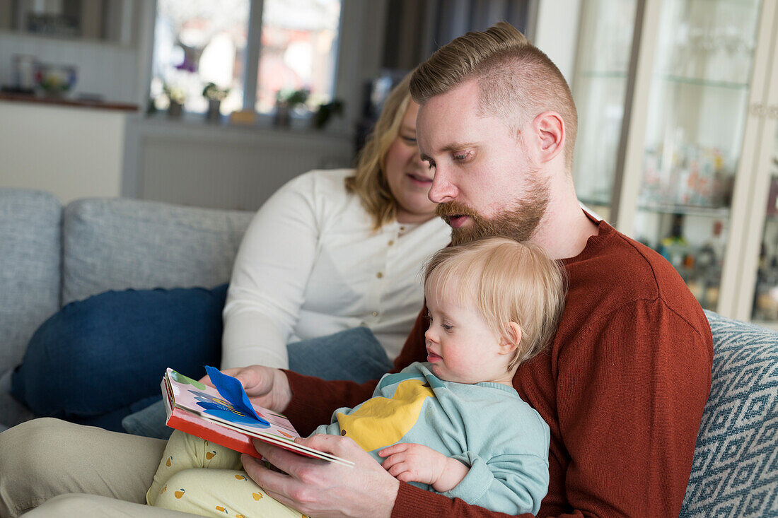 Eltern mit Baby mit Down-Syndrom lesen ein Buch auf dem Sofa