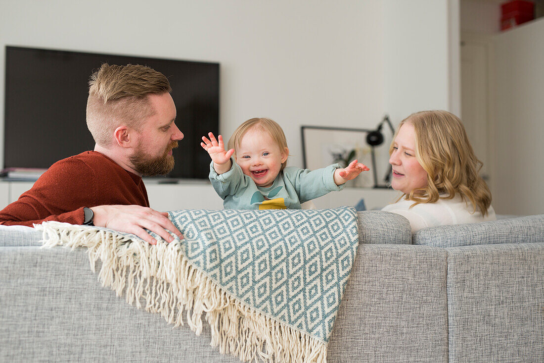 Eltern spielen mit lächelndem Baby Eltern mit Baby mit Down-Syndrom lesen ein Buch auf Sofa im Wohnzimmer