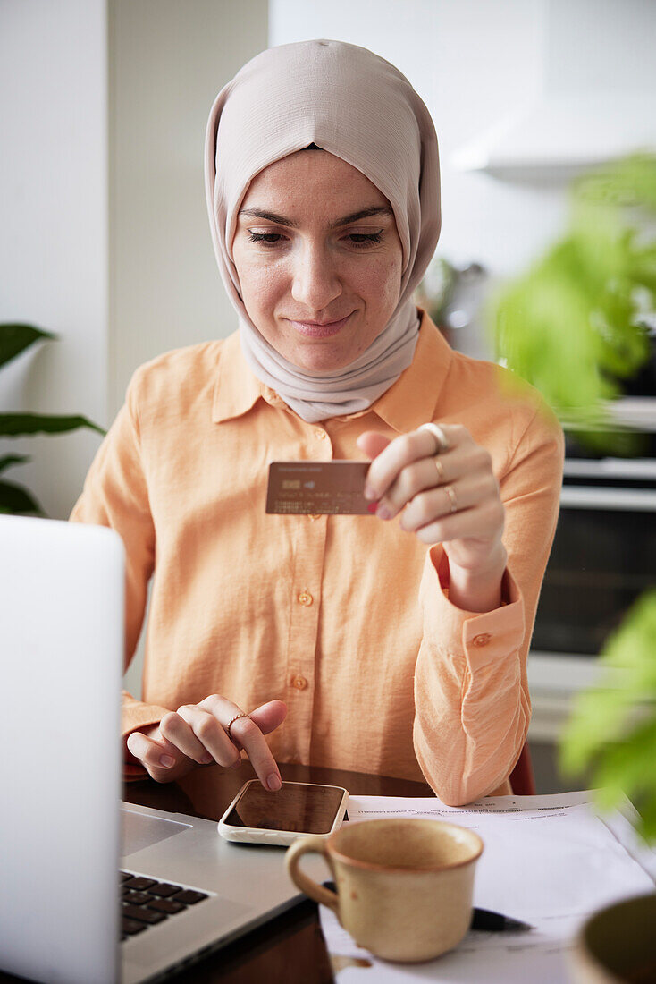 Smiling woman with hijab holding credit card while doing online shopping