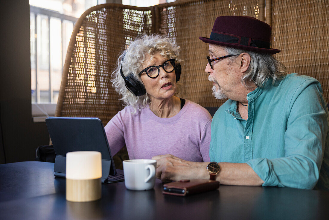 Senior couple sitting in cafe talking while working on digital tablet