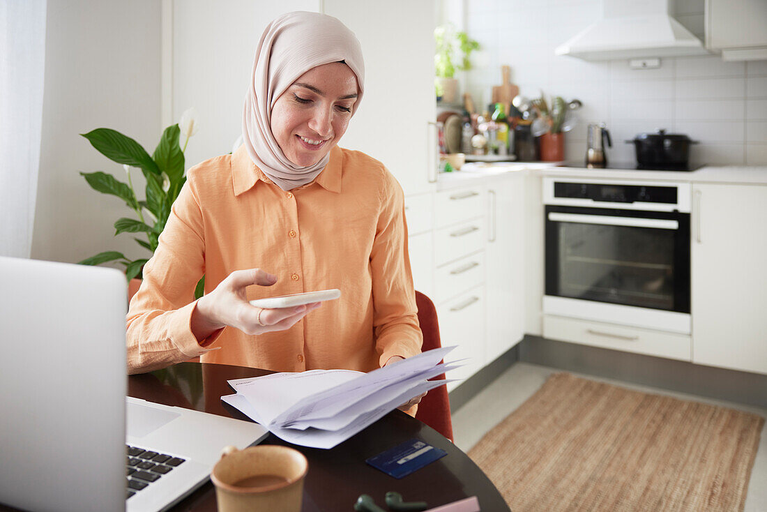 Lächelnde Frau mit Hijab, die ein Mobiltelefon benutzt und Rechnungen einscannt