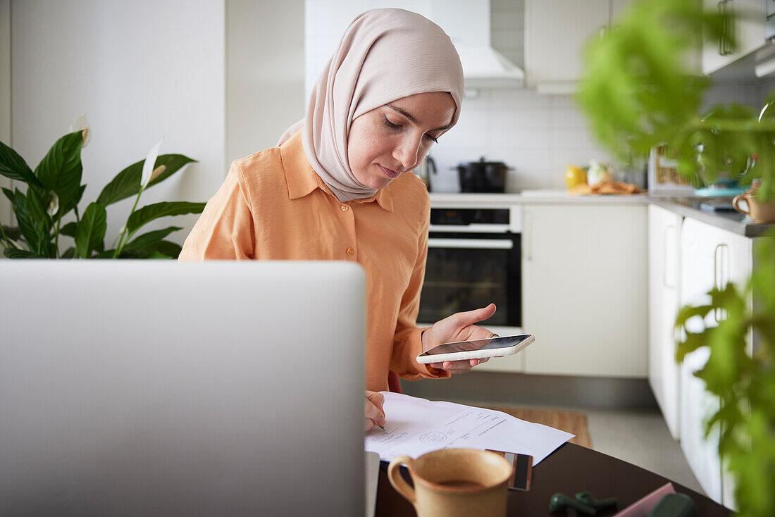 Lächelnde Frau mit Hijab benutzt Handy bei Heimarbeit in der Küche