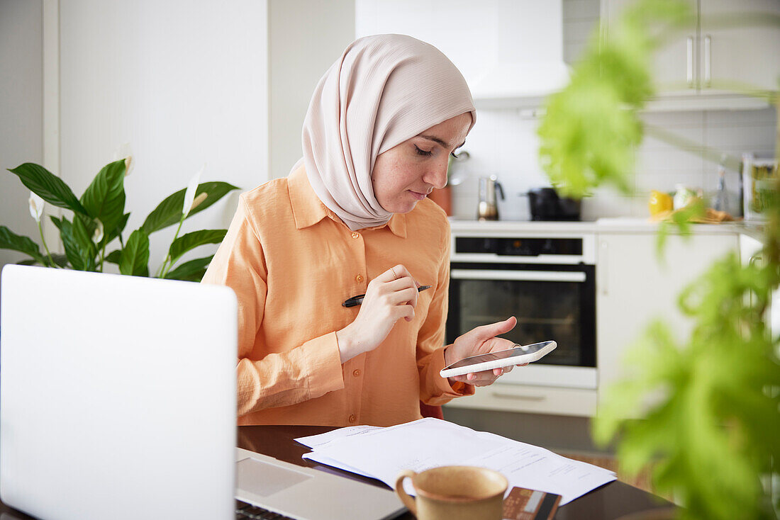 Lächelnde Frau mit Hijab, die während der Heimarbeit in der Küche telefoniert