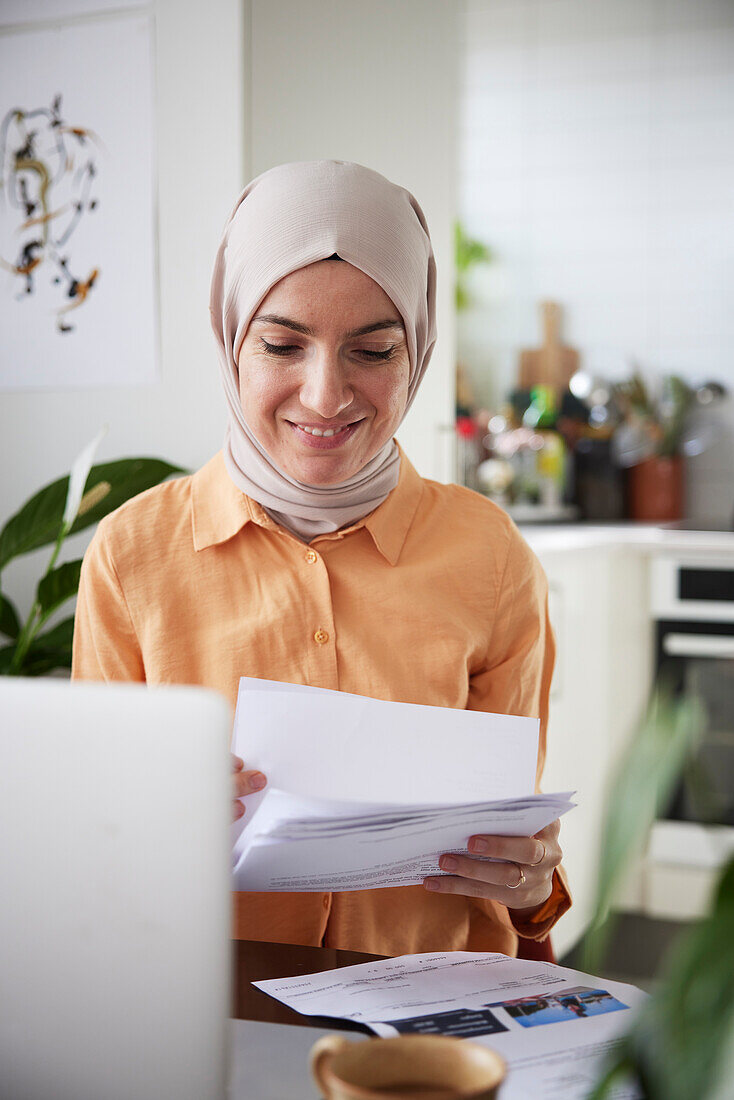 Lächelnde Frau mit Hidschab prüft Rechnungen zu Hause