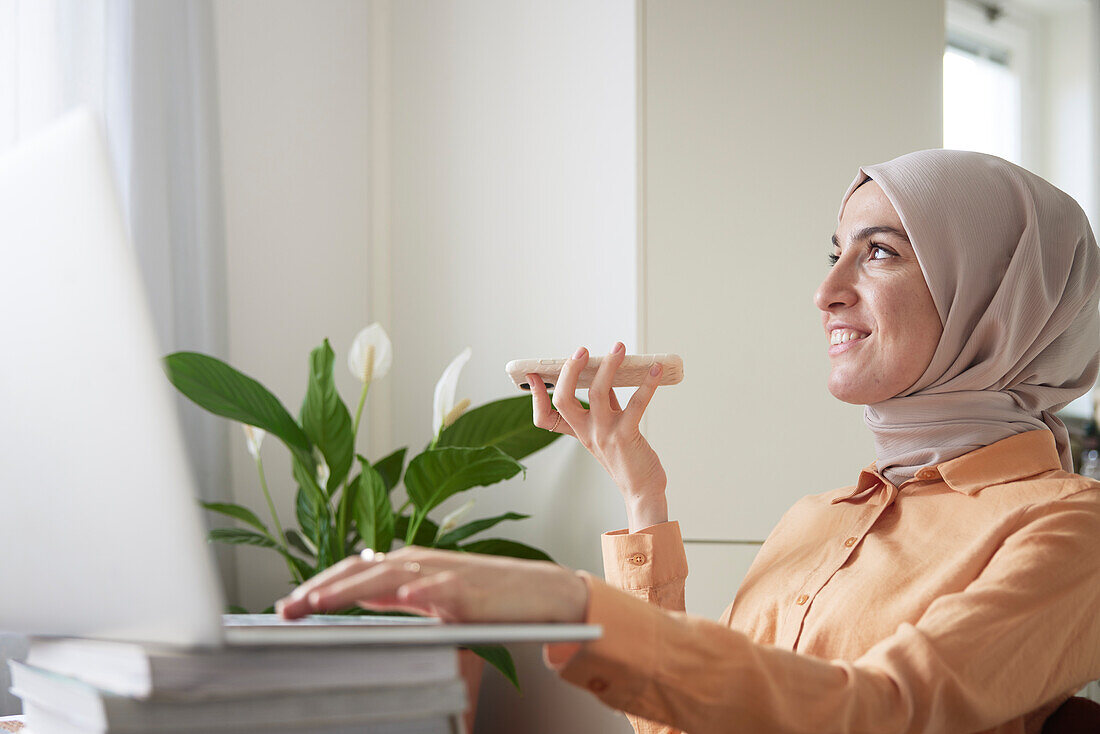 Lächelnde Frau mit Hijab unterhält sich im Lautsprechermodus mit ihrem Handy