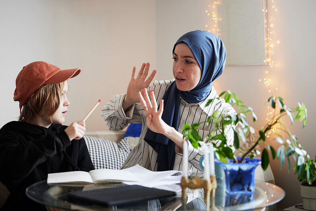 Mother wearing hijab holds up fingers while helping son doing math homework