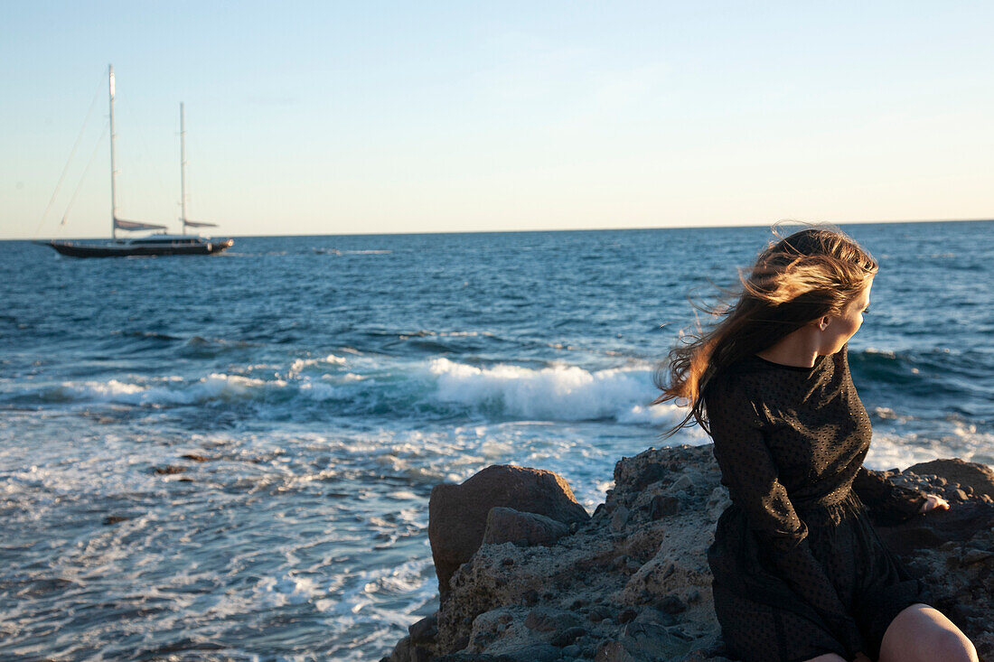 Woman sitting on rocks at sea with waives and sail boat in background