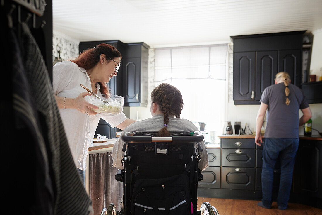 Eltern mit behinderter jugendlicher Tochter im Rollstuhl in der Küche, Mutter füttert Tochter im Rollstuhl während Vater Essen zubereitet