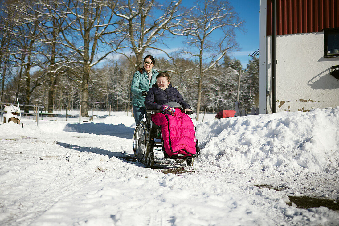 Mutter schiebt jugendliche Tochter im Rollstuhl bei Winterspaziergang