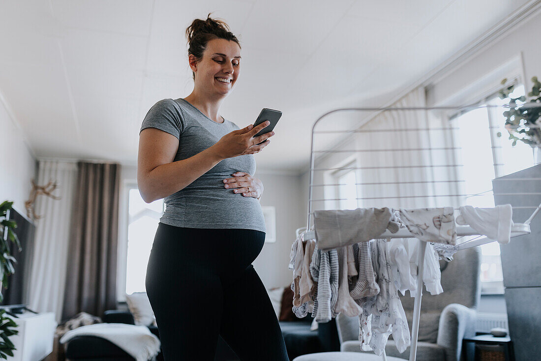 Schwangere Frau telefoniert bei der Hausarbeit und hängt Babykleidung auf den Wäscheständer