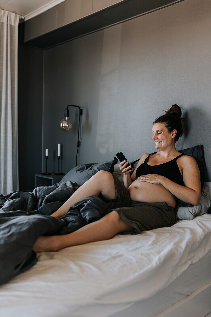 Glückliche schwangere Frau entspannt sich im Bett und benutzt das Telefon