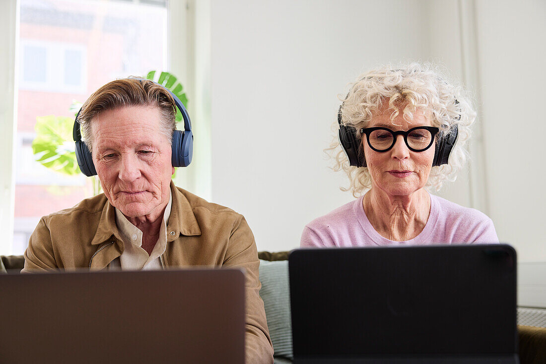 Senior man and woman sitting in living room and using laptop and digital tablet to edit podcast