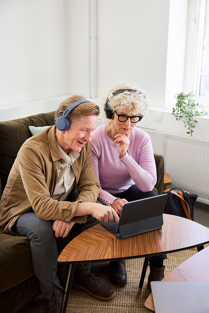 Älterer Mann und Frau sitzen im Wohnzimmer und benutzen Laptop und digitales Tablet, um Podcasts zu bearbeiten
