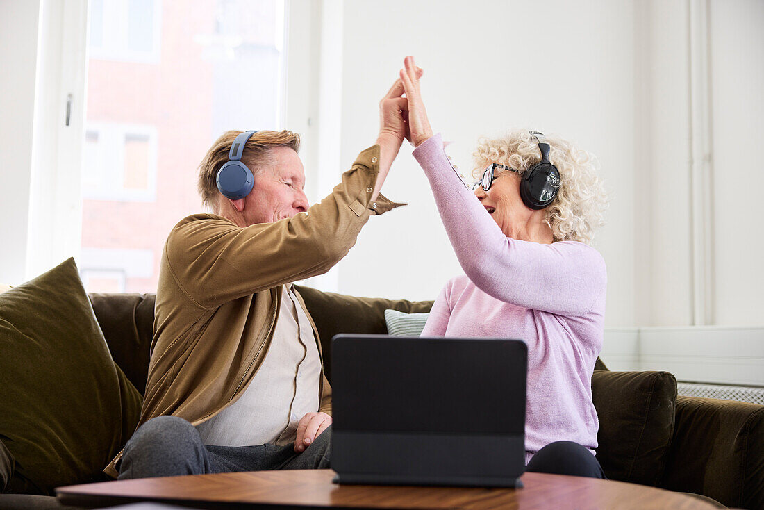 Älterer Mann und Frau geben sich gegenseitig High Five, während sie im Wohnzimmer vor einem digitalen Tablet sitzen