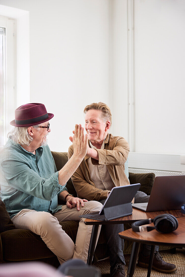 Senior men giving each other high five while sitting in living room in front of digital tablet