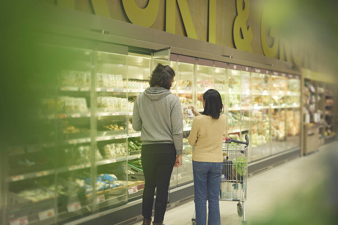 Rear vie of couple standing in front of fridges with vegetables while doing shopping in supermarket