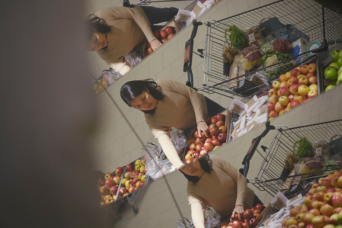 Frau mit Einkaufswagen wählt Äpfel aus und spiegelt sich im Supermarktspiegel