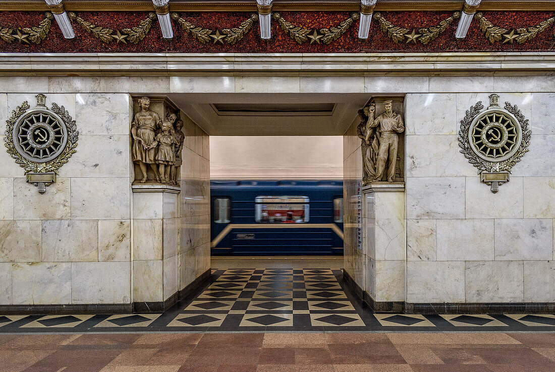 Narvskaya subway station in the city, built in the neoclassical style lined in white marble with many bronze inserts and a decorative frieze, a train moving past a platform. 