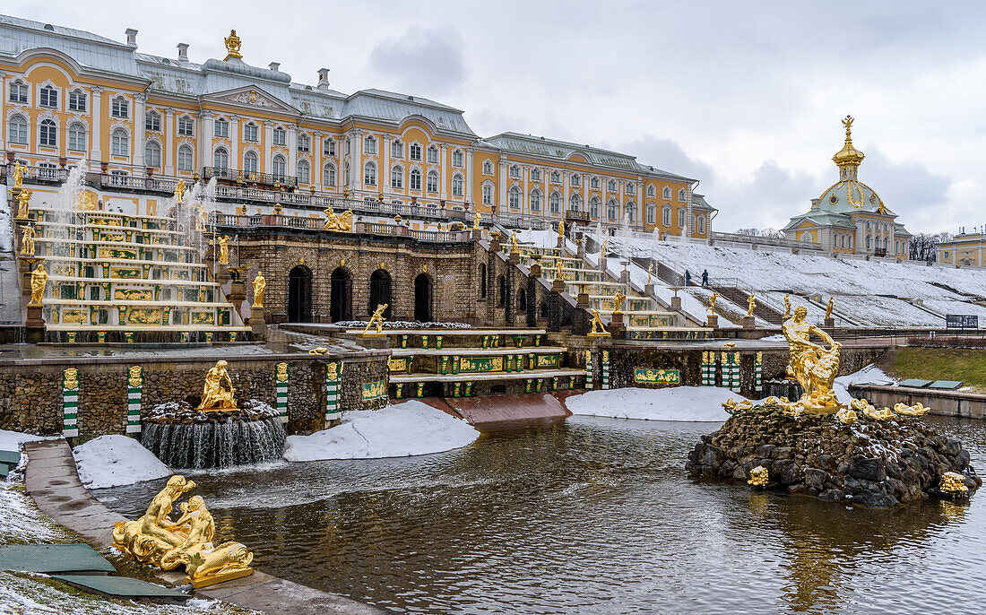 Die Fassade des Peterhofer Sommerpalastes der Romanow-Zaren, Springbrunnen in den Gärten und stilles klares Wasser, leichter Schnee auf Formschnittbüschen.