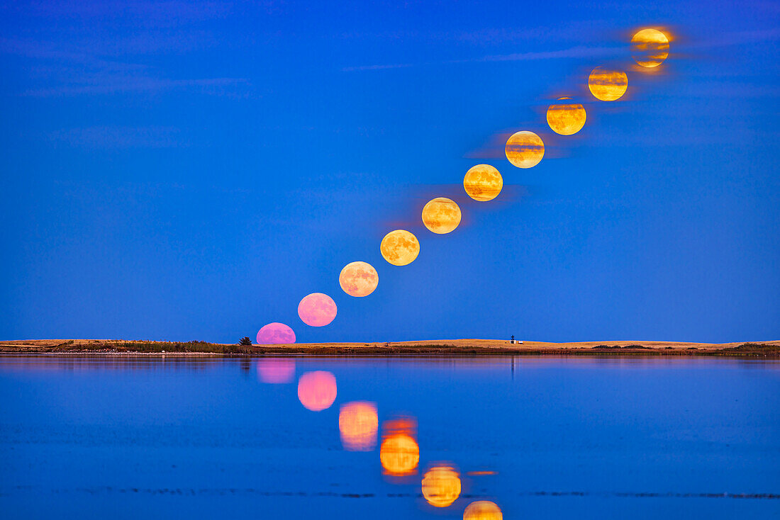 Der Aufgang des vollen "Jägermondes" vom 9. Oktober 2022, mit dem Mond, der sich in dieser Nacht im ruhigen Wasser des Crawling Lake Reservoirs im Süden Albertas spiegelt. Der Mond erschien sehr rosa, als er in den klaren Himmel in der Nähe des Horizonts aufstieg, und wurde dann gelber, als er in vereinzelte Wolkenbänder eintauchte.