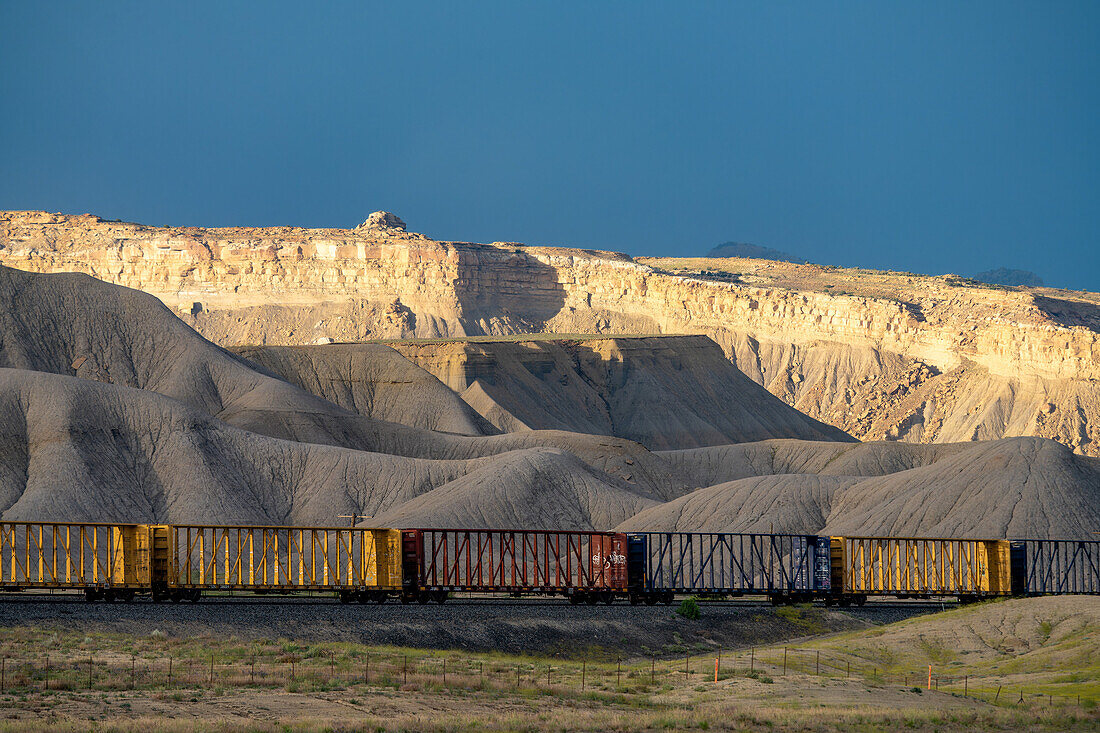 A line of centerbeam rail cars in the Green River Desert of Utah with spotlighting on the Book Cliffs behind.