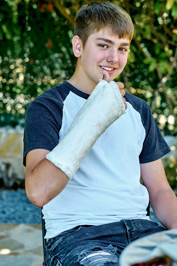 Porträt eines jungen kaukasischen Jungen mit gebrochenem und eingegipstem Arm, der auf einem Stuhl im Freien in einem Garten sitzt. Lebensstil-Konzept.