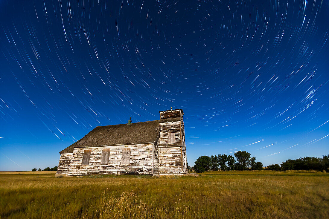 Zirkumpolarer Sternenschweif über der historischen, aber leider vernachlässigten St. Anthonys Church zwischen Bow Island und Etzikom, Alberta. Der Große Wagen ist links, Polaris oben. Die römisch-katholische Kirche wurde 1911 von englisch-russisch-deutschen Einwanderern erbaut. Sie diente einer schrumpfenden Gemeinde, bis sie 1991 geschlossen wurde. Damals fanden Arbeiter eine Zeitkapsel aus dem Jahr 1915 mit den Namen der damaligen Priester und Pfarrer. Im Sommer 2014 erlitt die Kirche ihre letzte Demütigung, als das Eisenkreuz auf ihrem Kirchturm gestohlen wurde. Es war noch da, als ich im M