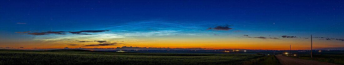 A panorama of a fairly bright display of noctilucent clouds to the northwest early in the evening on July 7, 2022. Even so, this was about 11:45 pm MDT. Capella is the bright star due north down the north-south range road at right. The bright NLCs contrast with the dark silhouettes of the closer and lower tropospheric clouds. A gibbous Moon lights the foreground.