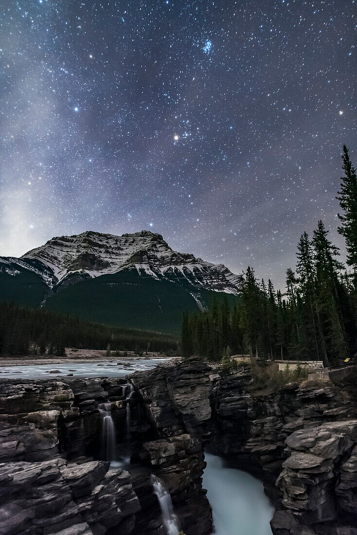 The Pleiades star cluster and the other stars of Taurus rising above Mount Kerkeslin at Athabasca Falls, in Jasper National Park, Alberta, October 22, 2016. The sky is brightening with the rising waning Moon off frame at left. Some cloud adds star glows and hazy patches to the sky.