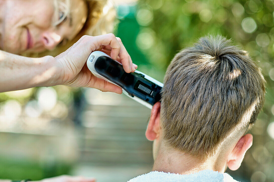 Porträt einer Mutter, die einem kleinen kaukasischen Jungen draußen im Garten die Haare schneidet. Lebensstil-Konzept.