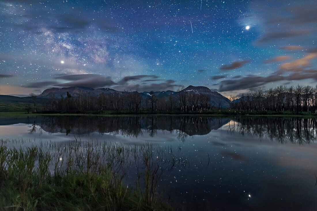 Jupiter (at right) and Saturn (at left) shining brightly in the sky and reflected in the still waters of Maskinonge Lake at Waterton Lakes National Park, Alberta, on June 17/18, 2018. The Milky Way is at left, Scorpius is at centre, and two satellite trails are at top. The sky is blue with solstice twilight. The trees on the opposite shore are charred from the Kenow Fire in September 2017. In the distance are Sofa Mountain and Viny Peak.