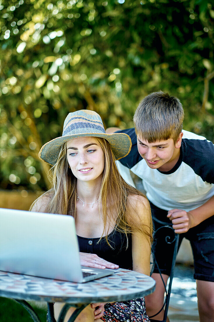 Portrait of a young caucasian woman with her younger brother posing outdoor in a garden with a laptop looking information in internet. Lifestyle concept.