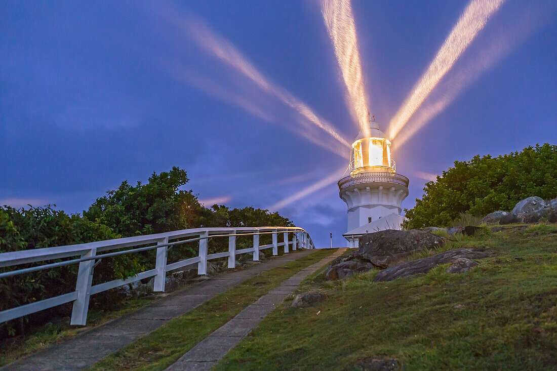 Der Leuchtturm von Smoky Cape mit seinen Strahlen in der bewölkten Abenddämmerung, die im leichten Regen leuchten. Er befindet sich in der Nähe von South West Rocks an der mittleren Nordküste von New South Wales, Australien. Der 1891 erbaute Leuchtturm war bis in die 1980er Jahre besetzt, als alle Leuchttürme automatisiert wurden. Der Leuchtturm wurde 1962 von Kerosin auf Elektrizität umgestellt und die Leuchtkraft auf 1.000.000 Candela erhöht. Der Leuchtturm verfügt über ein katadioptrisches Objektiv der Gebrüder Chance von 4 m Länge und 9 Feldern mit einer Länge von 920 m.