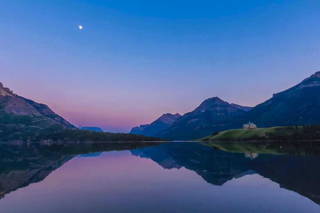 The waxing gibbous Moon over Upper Waterton Lake in Waterton Lakes National Park, Alberta with the iconic Prince of Wales Hotel in the distance, on a calm evening with still waters, rare in Waterton. This is an HDR stack of 3 exposures with the Canon 60Da and 16-35mm lens, shot from Driftwood Beach.