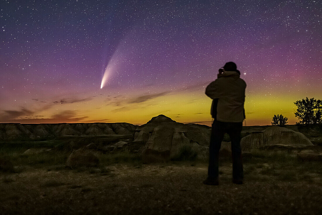 Ein Selfie bei der Beobachtung des Kometen NEOWISE (C/2020 F3) mit einem Fernglas in der dunklen, mondlosen Nacht vom 14. auf den 15. Juli 2020 vom Dinosaur Provincial Park, Alberta. Ein schwaches Polarlicht färbt den Himmel grün und magenta. Der schwache blaue Ionenschweif des Kometen ist zusätzlich zu seinem helleren Staubschweif sichtbar. Der Boden wird nur vom Sternen- und Auroralicht beleuchtet.