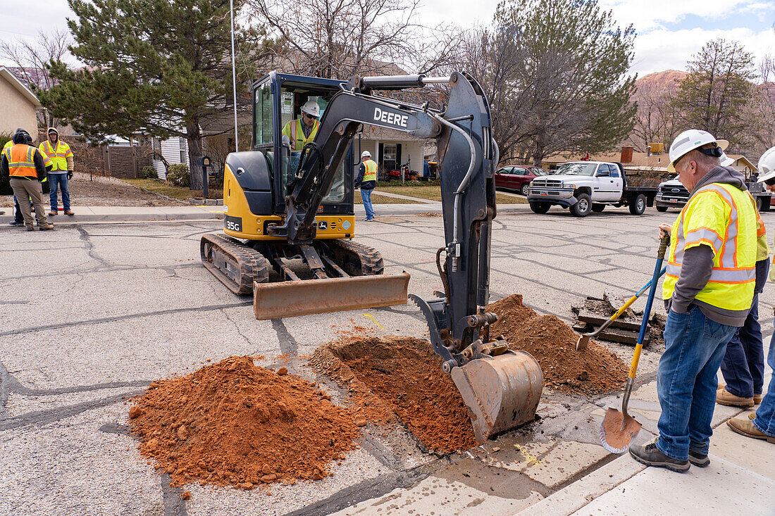A utility worker uses a track hoe to excavate a hole in the street to repair a utility line in a neighborhood.