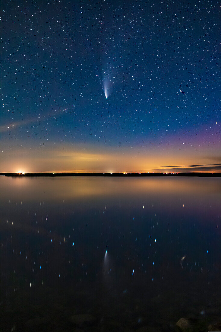 Comet NEOWISE (C/2020 F3) reflected in the still waters this night of Crawling Lake in southern Alberta. A dim aurora at right colours the sky magenta. Lingering twilight colours the sky blue. A meteor or more likely a flaring satellite appears at right and is also reflected in the water. Even in this short exposure, the two tails — dust and ion — are visible. This was July 20, 2020.