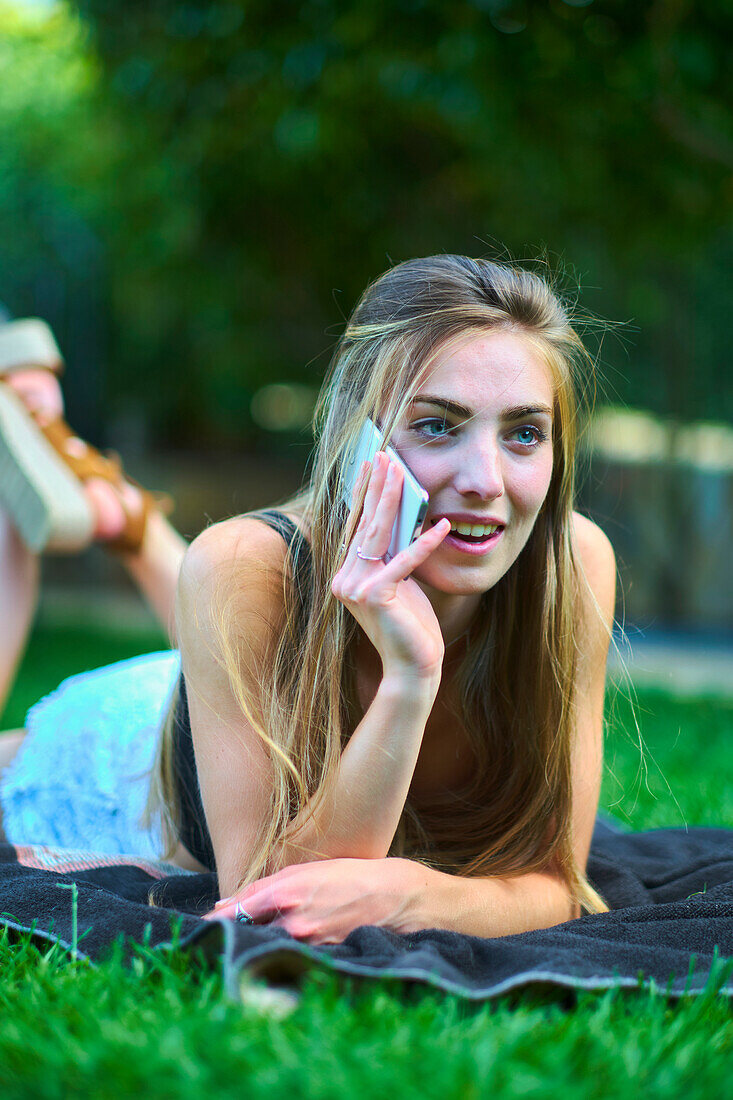 Porträt einer jungen schönen kaukasischen Frau in den 20ern, die in einem Garten im Gras liegt und mit ihrem Handy telefoniert. Lifestyle-Konzept.