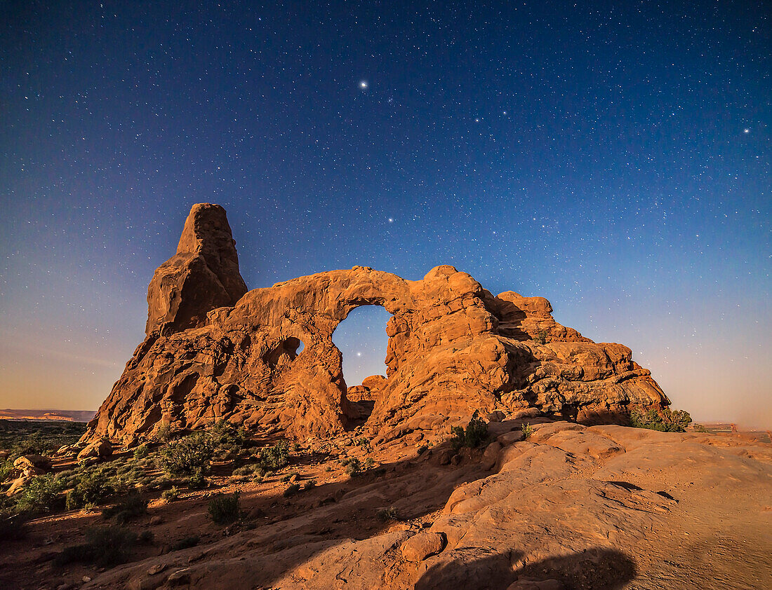 Eine mondbeschienene Szene am Turret Arch im Arches National Park, Utah, mit Sirius, der durch den Bogen scheint, Procyon über dem Bogen und darüber, oben in der Mitte, Jupiter, in der Nähe des Beehive-Sternhaufens. Die Sterne der Zwillinge befinden sich rechts von Jupiter. Der aufsteigende abnehmende Gibbous-Mond sorgt für die Beleuchtung.