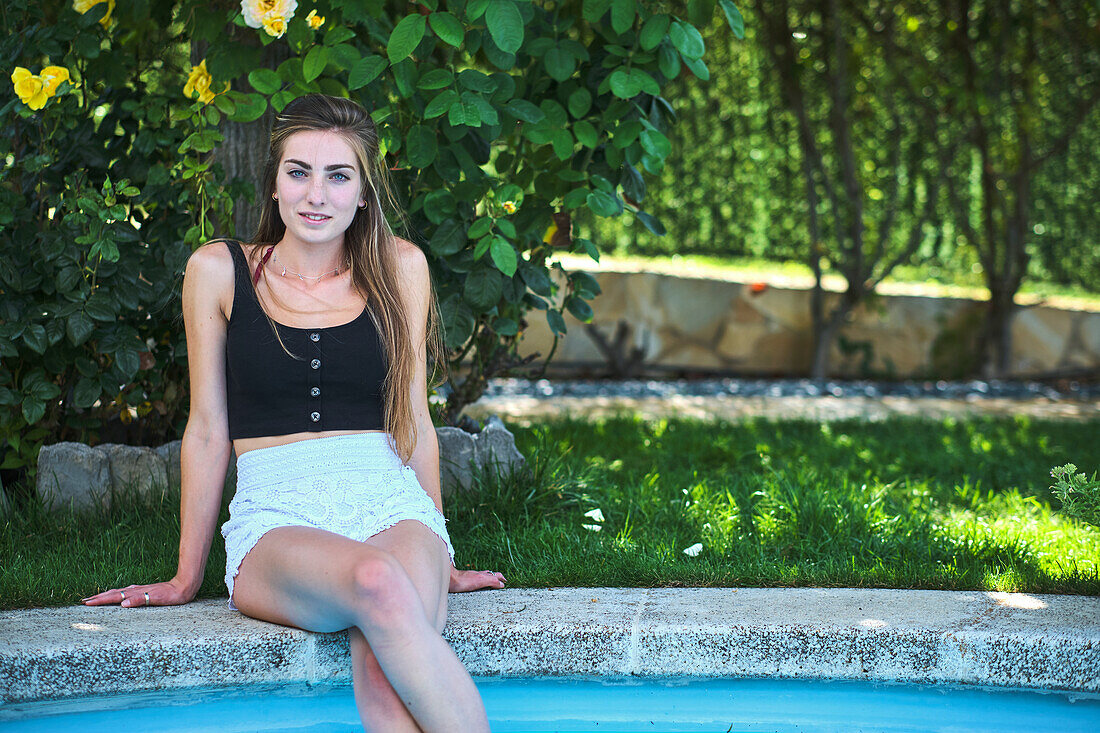 Portrait of a young beautiful caucasian woman in her 20´s with long hair and blue eyes sitting outdoor on the edge of a swimming pool. Lifestyle concept.